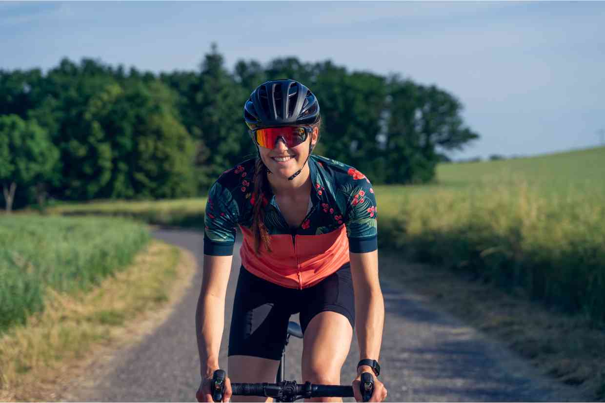 Mulher sorridente pedalando em estrada rural usando capacete MIPS preto e óculos espelhados.