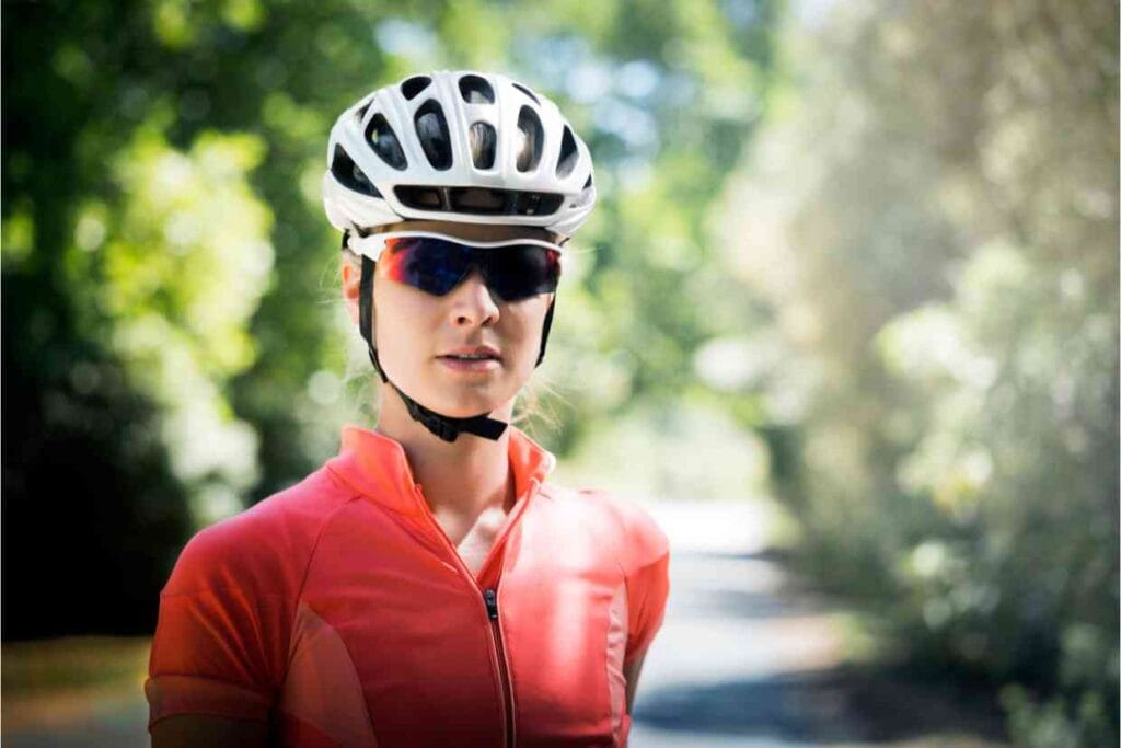 Ciclista mulher com capacete MIPS branco e óculos escuros em ambiente arborizado.
