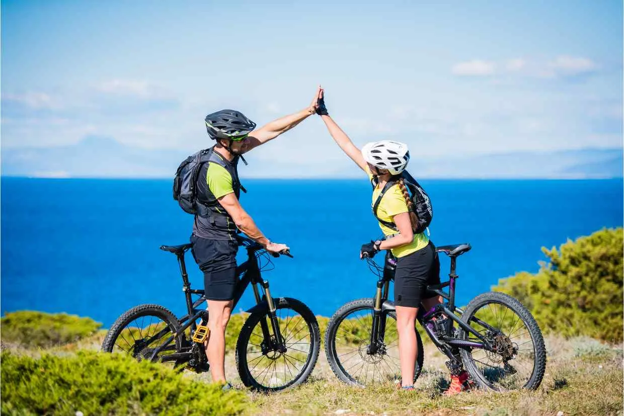 Imagem de dois ciclistas confraternizando no topo de uma trilha com uma bela vista do oceano ao fundo, representando um exemplo das vantagens das boas parcerias criadas.
