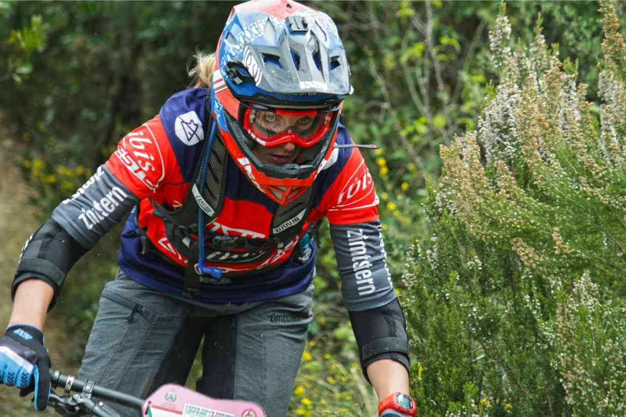 Mulher ciclista em competição, equipada com capacete downhill vermelho e óculos de proteção.