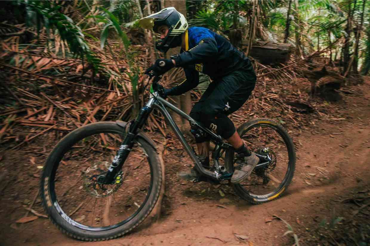 Ciclista contornando curva fechada em floresta, equipado com capacete downhill amarelo.