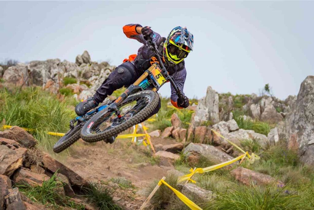 Ciclista de downhill com capacete downhill colorido fazendo curva em trilha rochosa.