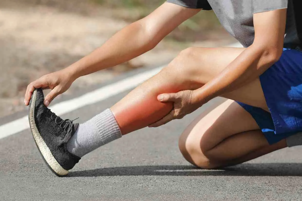 Ciclista sentado no chão com dor visível na panturrilha, região avermelhada indicando fadiga muscular.