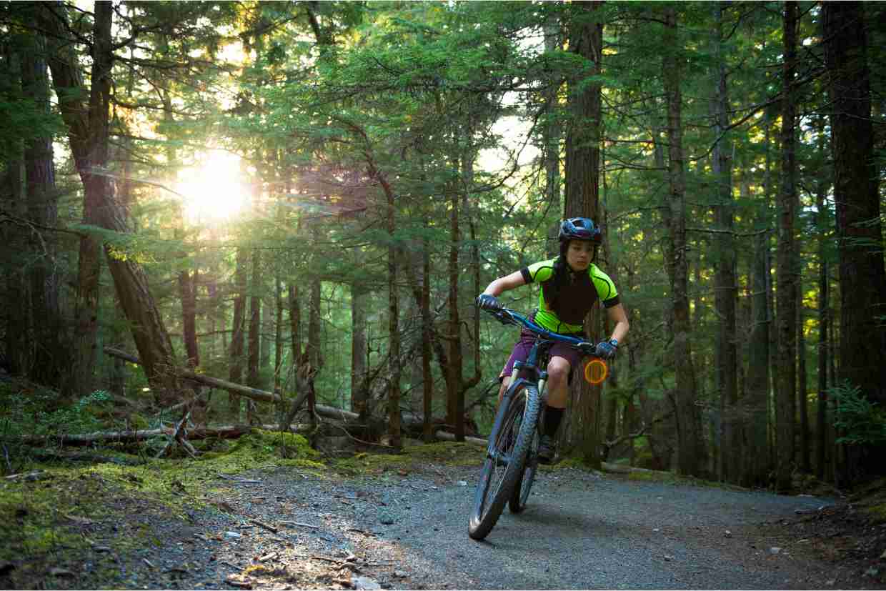Jovem ciclista em trilha de mountain bike cercada por árvores com luz do sol ao fundo.