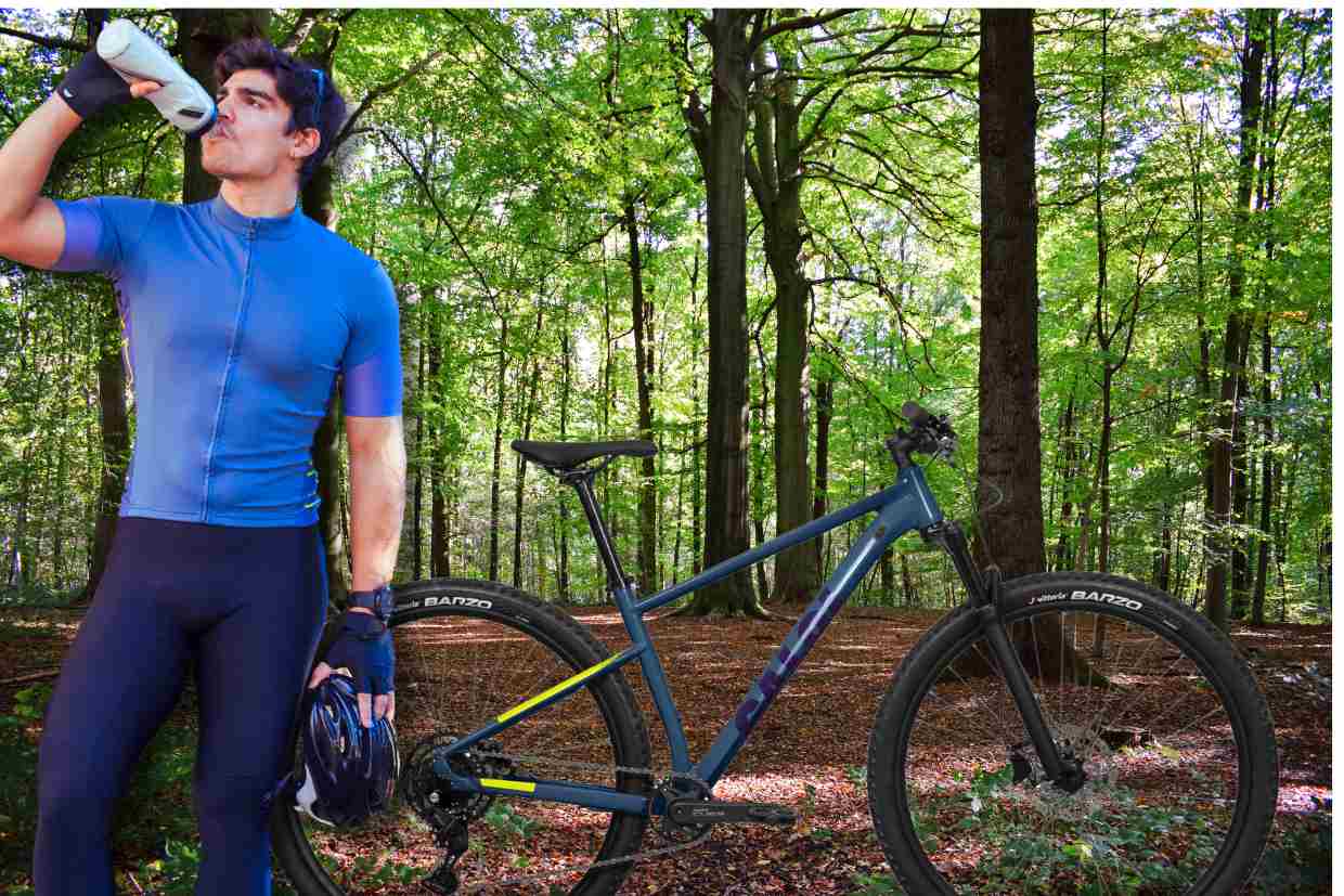 Ciclista em trajes esportivos tomando água ao lado da Bicicleta Caloi Explorer Comp azul em meio à floresta.