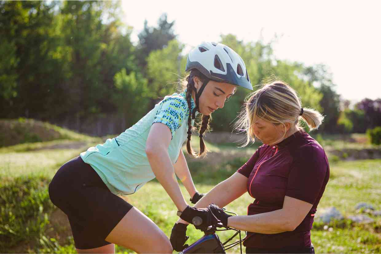 Duas mulheres ajustando a posição em uma bicicleta em um campo ao ar livre, se preparando para pedalar.