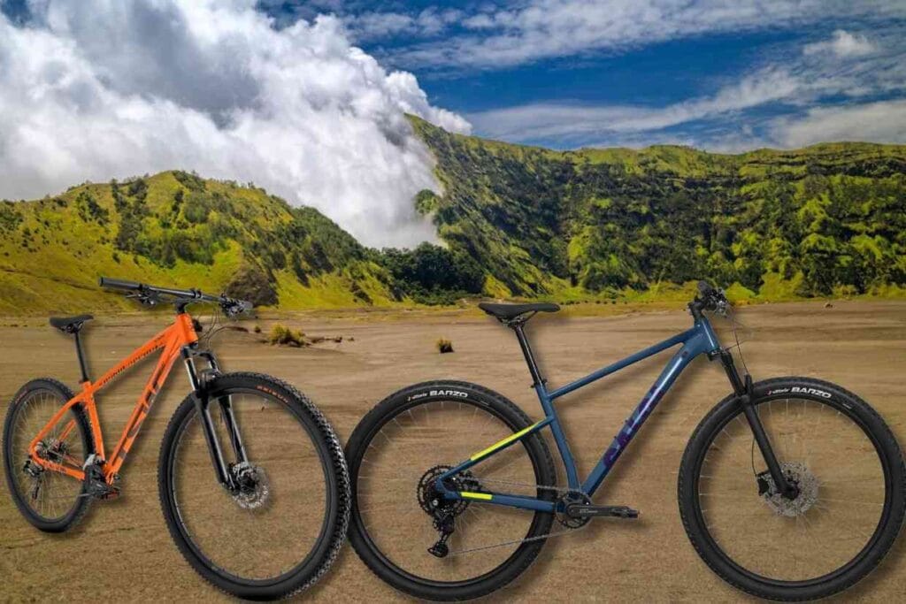 Imagem das bicicletas de MTB Caloi EXPLORER COMP SL e da bike TSW Hunch 16V paradas numa área de terra com uma montanha arborizada ao fundo.