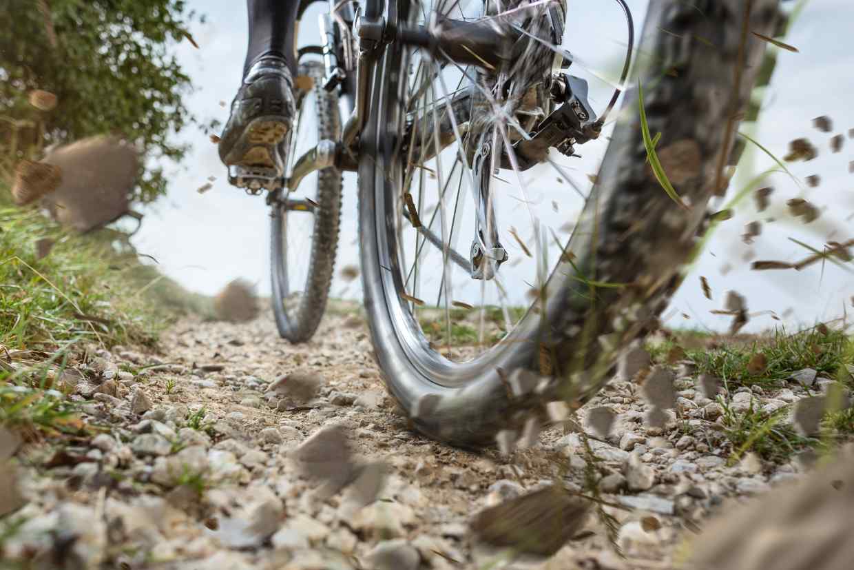 Ciclista passando com sua bicicleta MTB por uma trilha de cascalho, com algumas pedras soltas sendo lançadas a medida que a roda da bike passa por elas. Imagem: Canva.