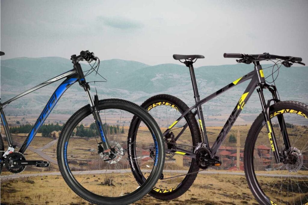 Duas bicicletas de mountain bike para iniciantes em primeiro plano: Oggi Big wheel 7.0 e Sense Fun Evo, tendo como fundo uma cidade rural distante e abaixo delas.