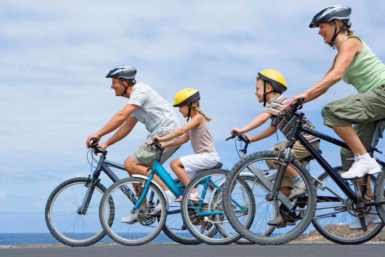 Família com dois adultos e duas crianças andando de bicicleta em estrada aberta beirando a orla, com o mar ao fundo usando diferentes modelos e tamanhos de bicicletas.