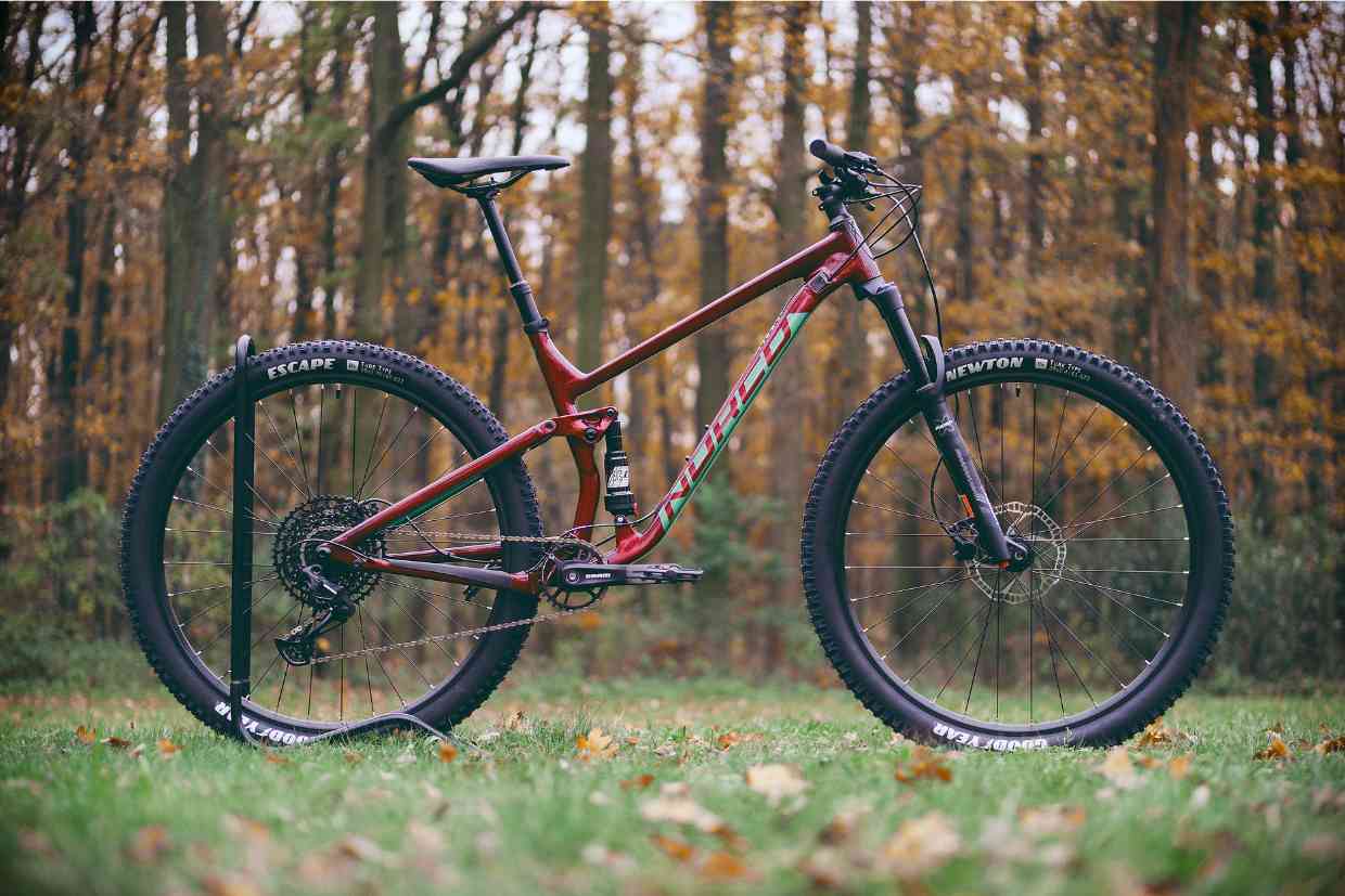 Bicicleta aro 29 vermelha posicionada em pé em frente a uma floresta no outono. Imagem mostra a importância do uso do adequado tamanho de bicicleta na prática do MTB.