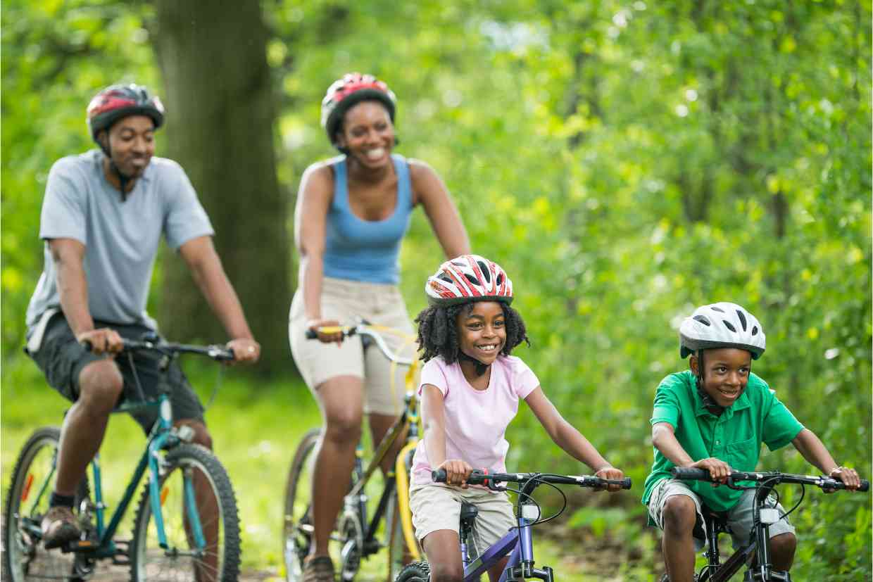 Família pedalando em trilha arborizada, com crianças e adultos sorrindo usando variados modelo e tamanho de bicicletas.