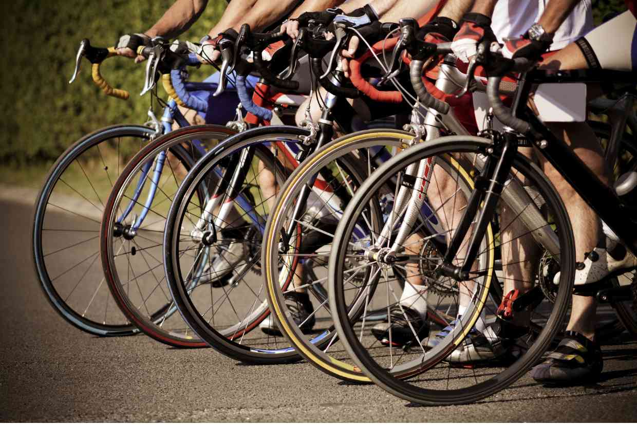 Linha de ciclistas em bicicletas de estrada prontas para largada em competição, nas suas bicicletas de modeloe e tamanhos diversos.