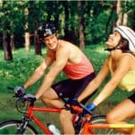 Casal pedalando juntos em área verde, sorrindo e aproveitando o passeio com suas bicicletas de tamanhos ajustados para maior segurança e conforto.