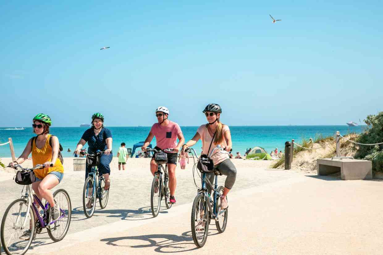 Grupo de pessoas pedalando na orla da praia em um dia ensolarado. Diferente tamanho de bicicleta para cada ciclista.