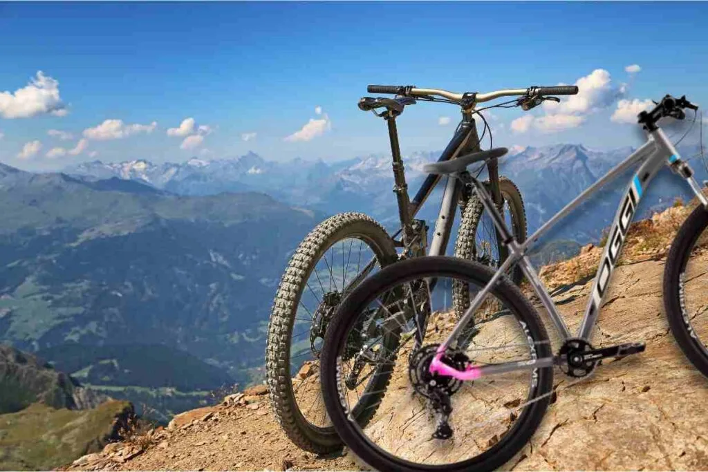 A bicicleta MTB Oggi Big Wheel 7.0 se destaca no topo de um penhasco com montanhas e o céu azul ao fundo. Imagem: Canva.