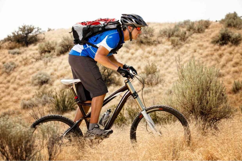 Ciclista com mochila pedalando uma bicicleta aro 29 em terreno seco e montanhoso.