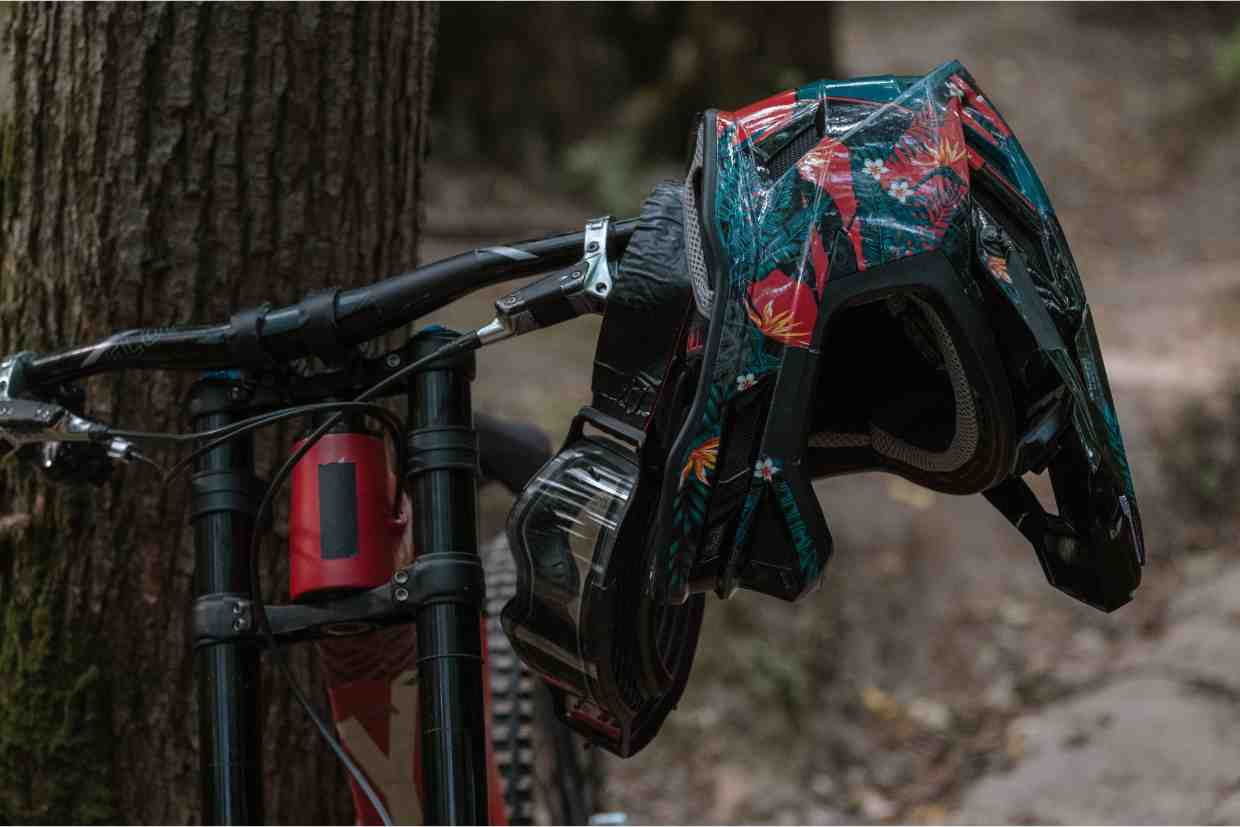 Capacete enduro MTB completo pendurado no guidão de bicicleta, com estampa camuflada, em ambiente de floresta.