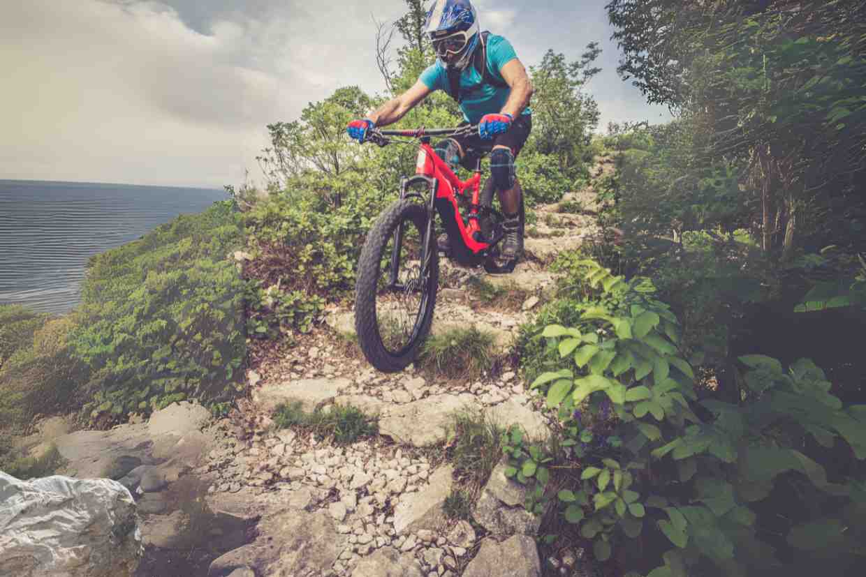 Atleta com capacete enduro MTB e luvas confrontando desníveis rochosos em trilha à beira do penhasco.