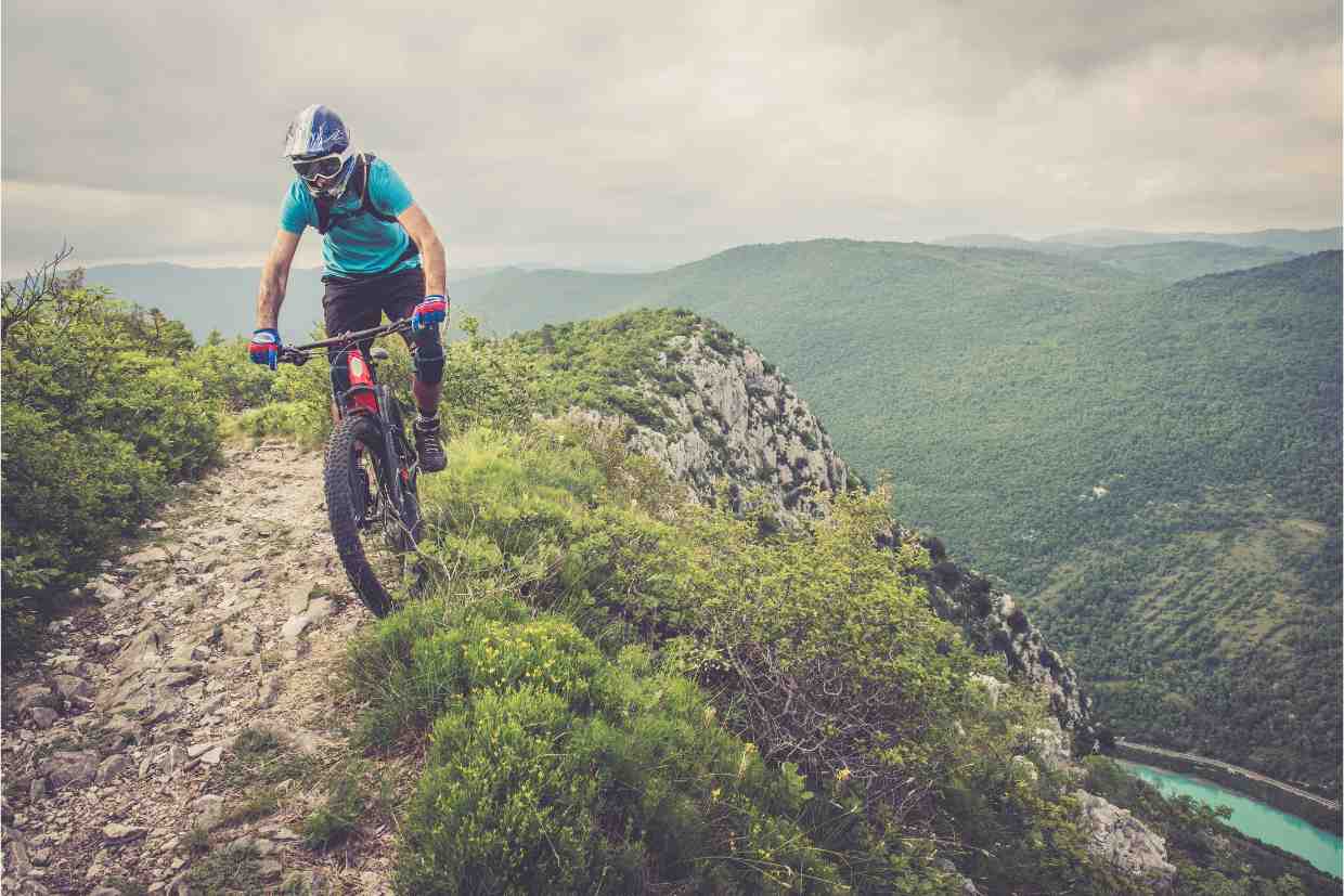 Ciclista usando capacete enduro MTB com queixeira completa, passando por trecho estreito e elevado com vista panorâmica.