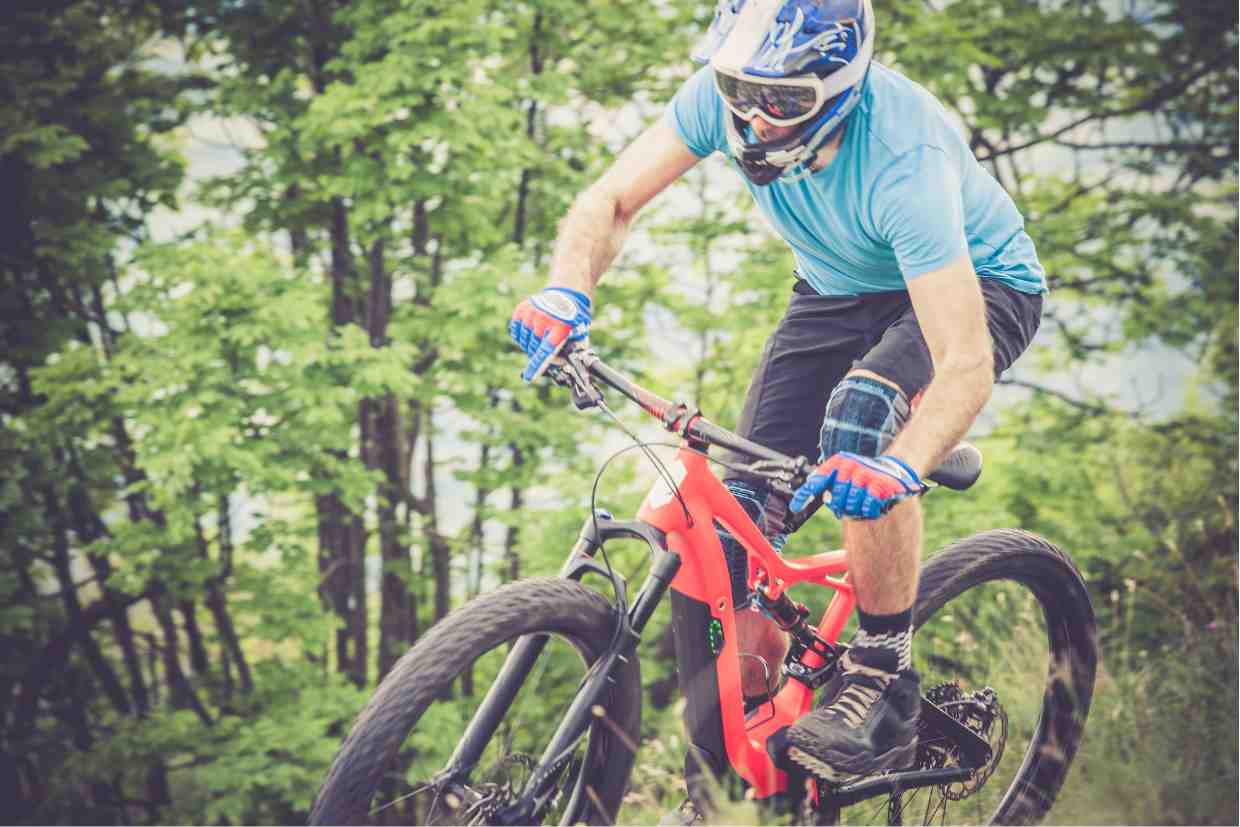 Close frontal de ciclista com capacete enduro MTB e óculos, focado na descida técnica na floresta.