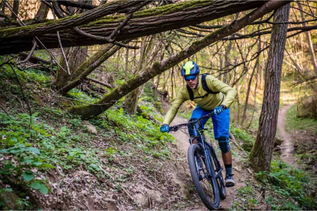 Ciclista em trilha arborizada usando capacete enduro MTB e viseira, descendo curva entre raízes e galhos.