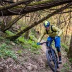 Ciclista em trilha arborizada usando capacete enduro MTB e viseira, descendo curva entre raízes e galhos.