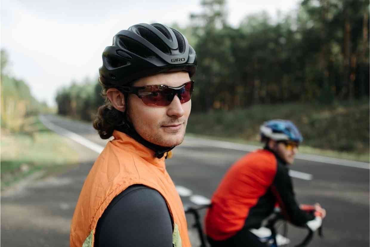 Imagem de dois ciclistas parados na beira da estrada, um deles usando o capacete de ciclismo Giro Cinder MIPS preto, aguardando para fazer a travessia da pista.