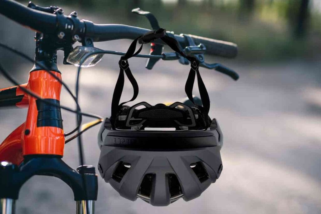 Capacete Giro Manifest Spherical preto pendurado por suas alças no guidão de uma bicicleta de MTB de cor laranja.