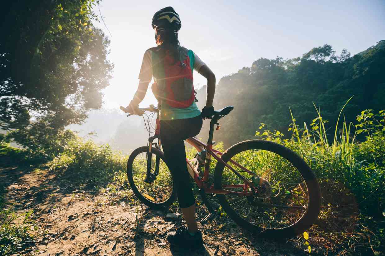Mulher de pé numa trilha, segurando sua bicicleta como o Capacete Giro Manifest Spherical preso a cabeça, enquanto observa o sol se pondo no fim da tarde.