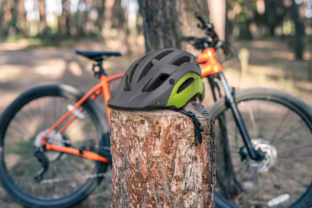 Capacete Giro Manifest Spherical colocado sobre o toco de uma árvore com uma bicicleta de MTB laranja estacionada ao fundo desfocado da imagem.
