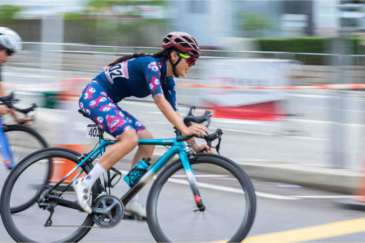 Ciclista em competição usando capacete bike feminino rosa e azul, focada e em movimento na estrada.