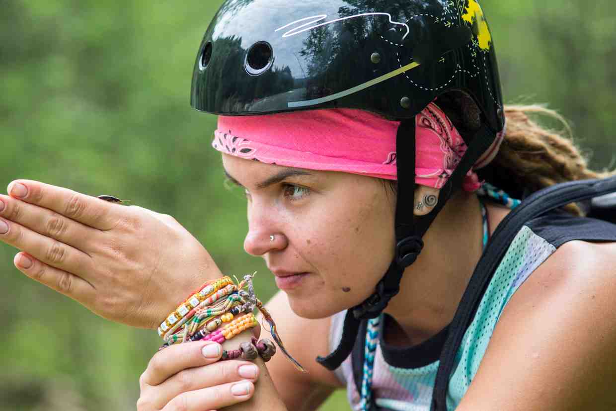 Mulher com capacete bike feminino estilo urbano e acessórios coloridos observa inseto em ambiente natural.
