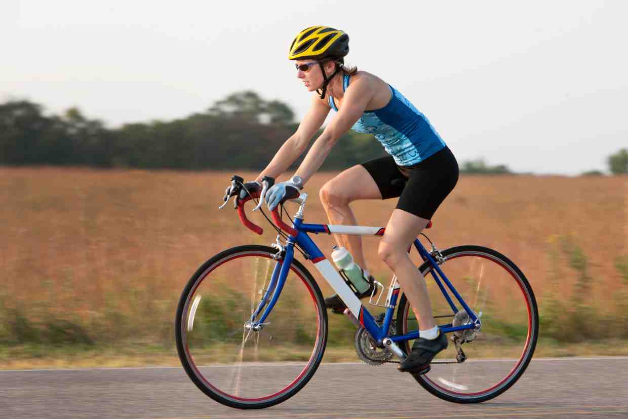 Mulher pedalando com capacete bike feminino amarelo em estrada plana, representando segurança e desempenho.