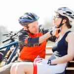 Homem ajudando ciclista a ajustar o capacete para MTB enquanto descansam no banco de uma praça.