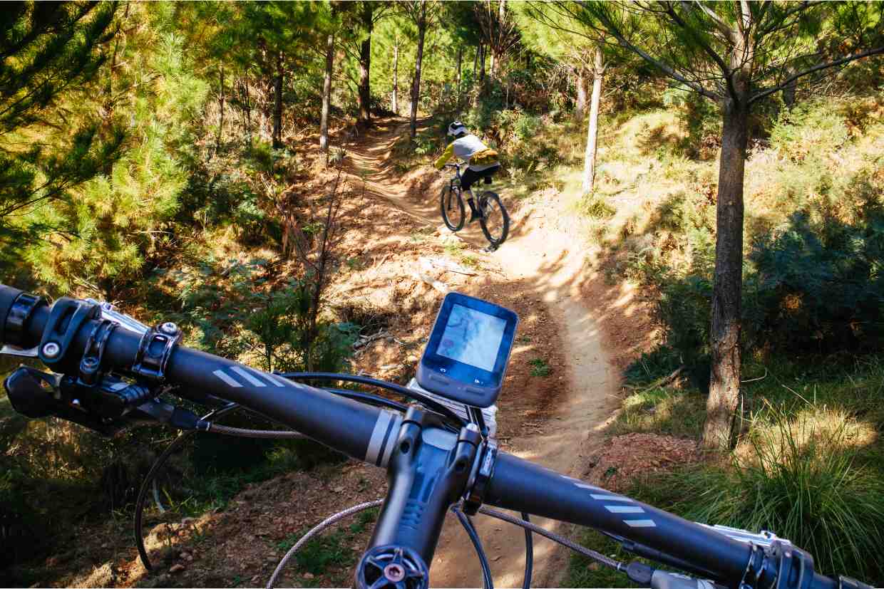Perspectiva do guidão da bike Groove Hype 70 em trilha de mata, com outro ciclista à frente no trajeto.