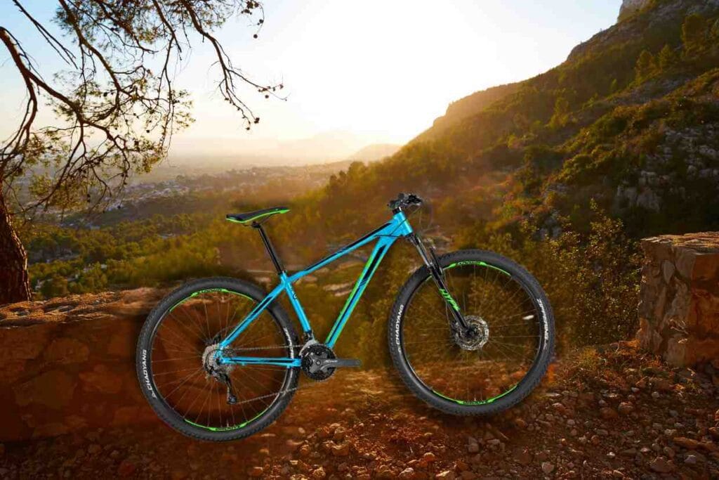 Bike Groove Hype 70 azul estacionada em uma trilha de montanha com vista panorâmica ao pôr do sol.