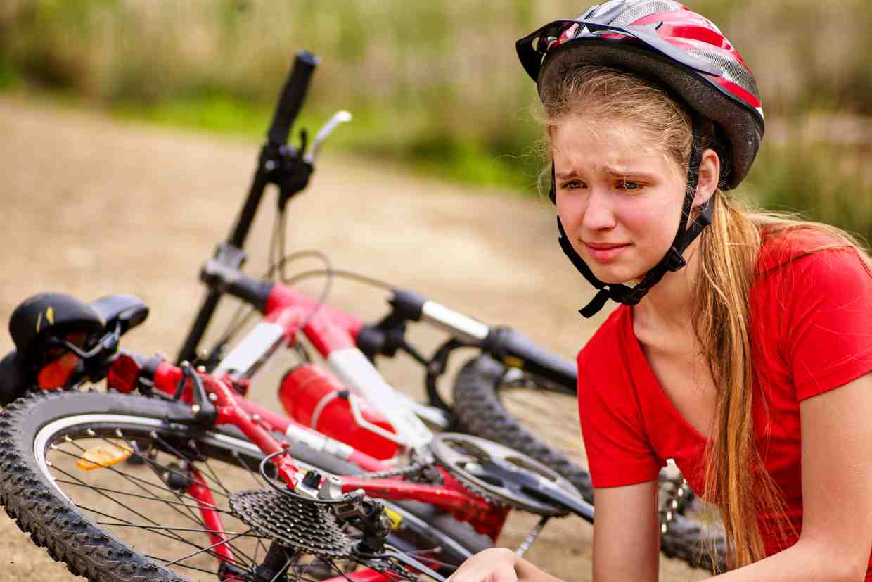 Jovem ciclista sentada próximo à bicicleta caída, com expressão de dor após uma queda usando capacete, ressaltando os riscos de usar um capacete fora da validade.