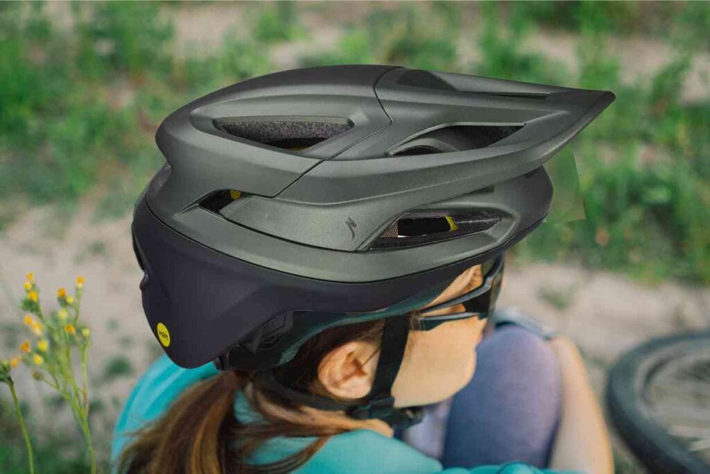 Jovem ciclista usando um capacete Specialized Camber cinza sentada no chão, com sua bike ao lado, enquanto observa a paisagem.