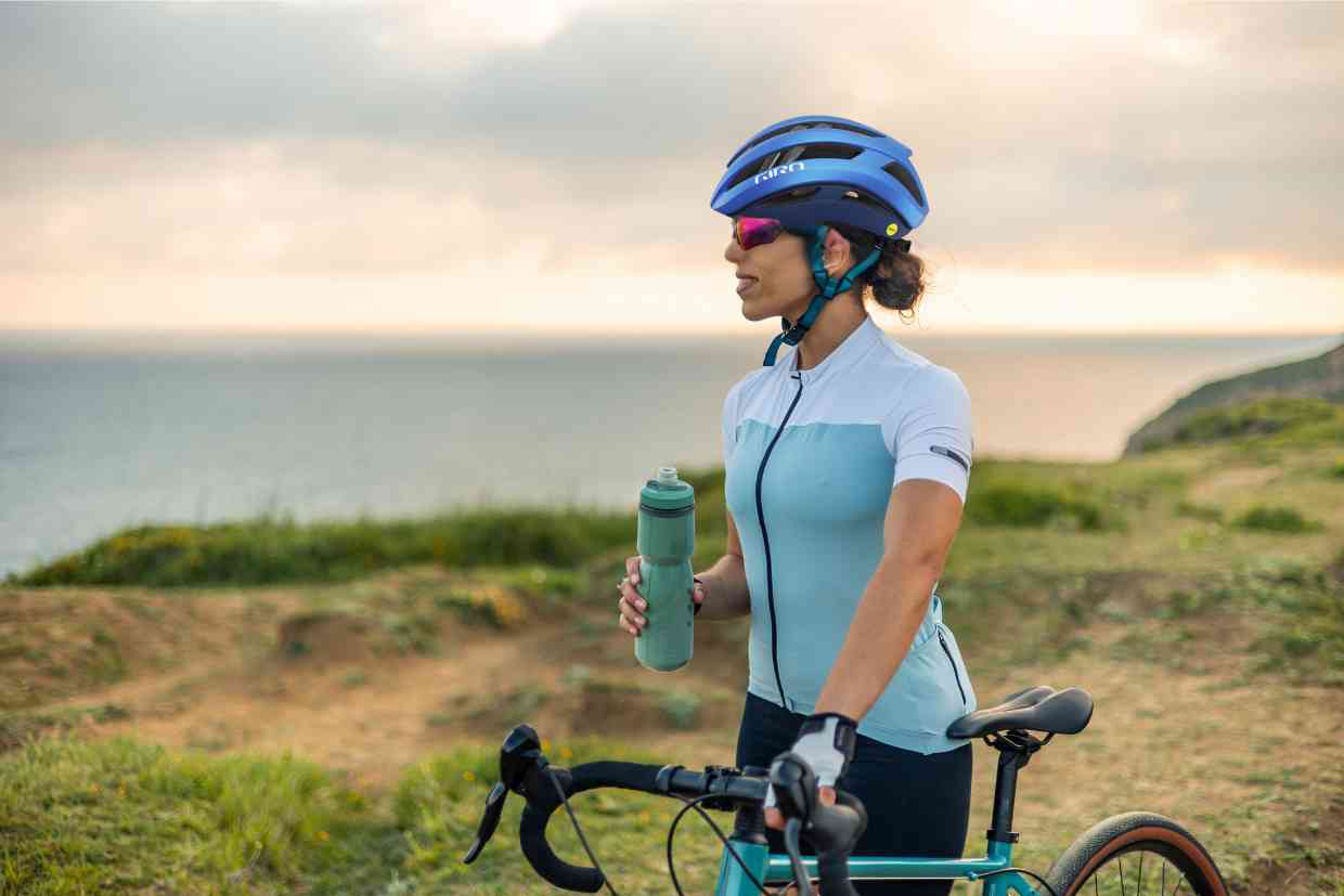Ciclista observa o mar enquanto segura sua bicicleta e uma garrafa de água, usando óculos de sol e capacete Giro Aries Spherical.