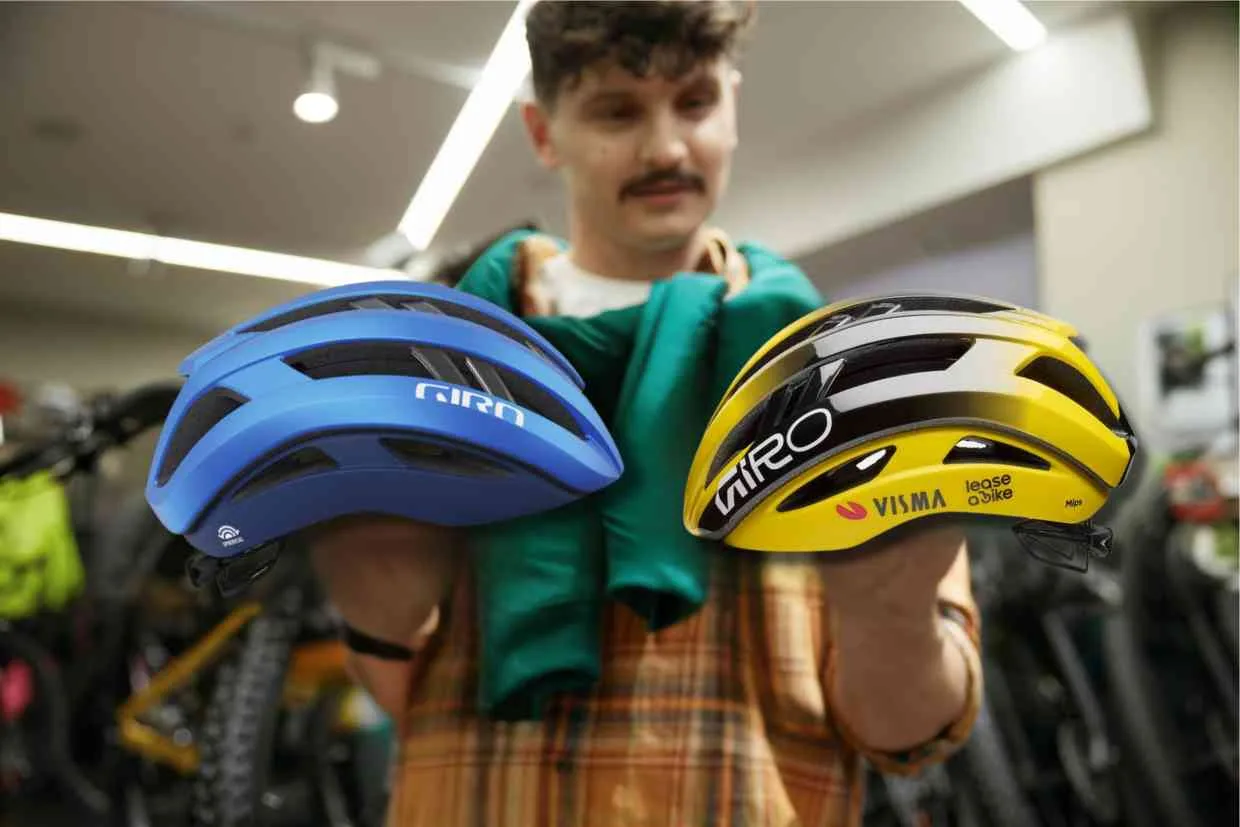 Vendedor de uma bicicletaria exibe dois capacetes Giro Aries Spherical um azul e outro amarelo a um cliente interessado.