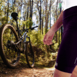 Ciclista de pé numa trilha mtb usando a bermuda Márcio May Light Bag, observando sua bike apoiada num arbusto.