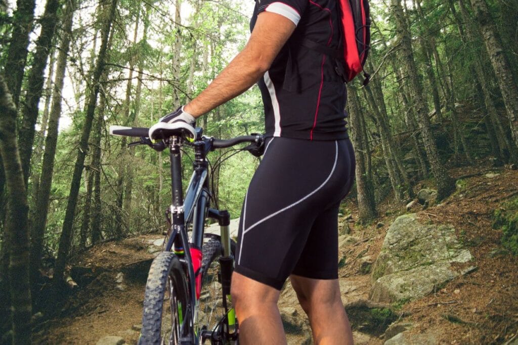 Ciclista segurando sua bike na entrada de uma trilha de muitas pedras, usando uma bermuda de ciclismo masculina preta.