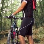 Ciclista segurando sua bike na entrada de uma trilha de muitas pedras, usando uma bermuda de ciclismo masculina preta.